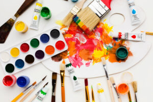 An overhead view of paintbrushes, paint palettes, and acrylic paint tubes that are scattered all over the place.